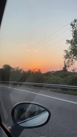 The video shows a view from inside a car looking out the passenger side window as the car travels down a road. The sun is setting, casting an orange glow in the sky above a line of trees. The road has a white stripe down the middle and a guardrail separates it from a grassy shoulder. The car's side mirror is visible in the lower part of the frame, reflecting the interior of the car. The sky is a pale blue transitioning to orange and pink near the horizon. There are some thin clouds visible in the sky. Overhead power lines are also visible.