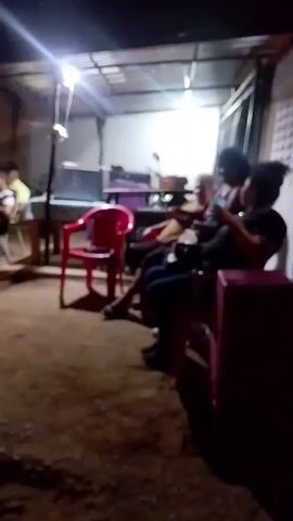 The video depicts an outdoor gathering at night, likely a party or social event. Initially, a few people are seated under a covered area illuminated by bright lights. There are red and pink chairs, and a table-like structure is visible. The camera then pans to reveal a larger group of people mingling in an open space. They are dressed casually, and some appear to be talking and gesturing. A sound system or speaker setup is visible in the background, suggesting the presence of music. The setting seems to be in a residential area. The overall atmosphere is relaxed and social.