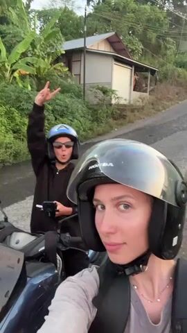 A video shows two people on a motorcycle. The person in the foreground, wearing a helmet, smiles at the camera. The person behind them, also wearing a helmet and sunglasses, makes a peace sign. They are on a road in a rural area with lush vegetation and palm trees. The sky is a gradient of blue and pink, suggesting it is either sunrise or sunset. Several other motorcycles are visible on the road ahead.