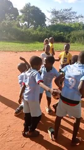 A group of children, mostly wearing matching light blue outfits, are playing on a red dirt field. They seem to be preparing for a game, possibly soccer, as one child holds a soccer ball. Some children are clapping, and another is running in the background. The environment includes trees, mountains in the distance, and a clear blue sky.