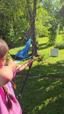 A person wearing a pink t-shirt with a braid tied with colorful ribbons is aiming a bow and arrow. They are in an outdoor setting with lush green grass, trees, and a blue hammock strung between two trees. In the background, there is a white target with an orange bullseye. The person's face is mostly obscured by their arm as they draw the bowstring. The sun is shining, casting shadows on the grass.