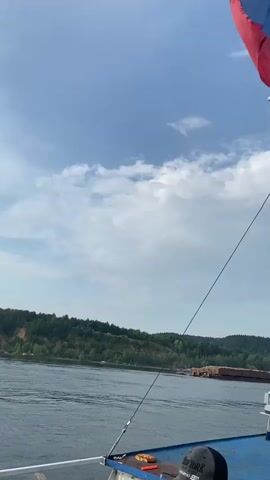 A partially visible Russian flag flies from a pole on a boat as it travels down a wide river. The camera is positioned on the deck of the boat, showing a section of the railing and the water. In the background, a lush green, tree-covered hillside lines the opposite bank of the river. Further in the distance, a barge filled with logs is visible moving down the river, and beyond that, another smaller vessel. In the foreground, three young individuals are visible on the deck of the boat, two boys and a young girl. One boy, wearing a black baseball cap, is looking down, while the other two are looking out at the scenery. The sky is a bright blue with scattered white clouds. The overall scene depicts a leisurely boat trip on a sunny day.