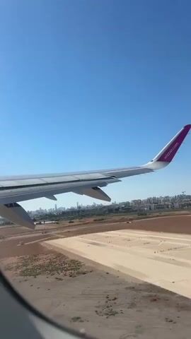 A view from an airplane window shows a city in the distance with buildings and structures. The airplane's wing, painted white with a purple tip and the logo "wizzair.com" is visible in the foreground, angled upwards. Below the wing, the ground consists of dry, sandy terrain with some patches of green vegetation and what appear to be agricultural fields or construction sites. The sky is a clear, bright blue. The perspective suggests the plane is either taking off or landing, with the ground appearing to recede or approach rapidly.