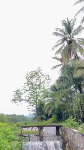 The video shows a small, artificial waterfall or dam in a tropical environment. The waterfall is constructed from concrete and consists of three tiers, with water cascading over each level. The surrounding landscape is lush, with various trees and vegetation visible, including tall palm trees. A simple bridge or walkway spans the top of the waterfall structure. The sky is overcast, and the overall atmosphere is tranquil and scenic.