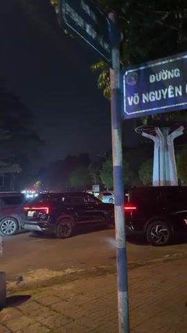The video captures a night scene on Võ Nguyên Giáp Street in Măng Đen, Vietnam. Cars are seen in moderate traffic, with headlights and taillights illuminating the road. Several black SUVs are visible on the street, and a white SUV (resembling a VinFast VF 3) is either parked or slowly moving on the side. A woman wearing a hat and jacket walks on the sidewalk next to a tree. Streetlights cast a warm glow, and trees line the road, indicating an urban or suburban setting. The street sign for "ĐƯỜNG VÕ NGUYÊN GIÁP" is prominently displayed at the beginning of the video.