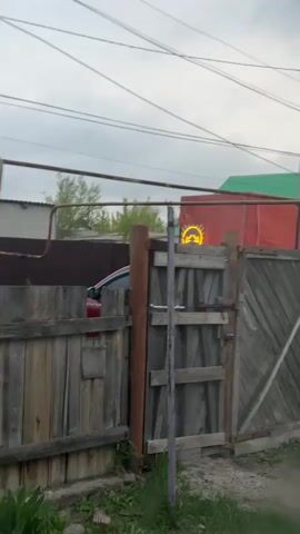 The video shows an outdoor scene, possibly in Aksay, Kazakhstan, on an overcast day. In the foreground, there is an old wooden fence and a closed wooden gate with some rusty metal hardware. Behind the fence, a red car is partially visible on the left, with some faint white text ("CTEPWA" or similar) on its side. Directly behind the gate, an illuminated yellow circular sign with a blue and white display changes its imagery.

Initially, the sign displays a left arrow with a smaller arrow pointing up and right, suggesting a two-way or merging traffic situation. It then changes to a symbol resembling a car driving over a bump or a hill, indicating an uneven road or speed bump. Finally, the sign displays a single left arrow within a blue circle, likely indicating a mandatory left turn or "keep left."

In the background, a building with a bright green roof is visible, along with a large red tarp or banner. Overhead power lines crisscross the sky, and a street light pole is visible on the right.