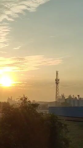The video is a time-lapse recording of a sunrise over an industrial area. The sun is visible on the left side of the frame, casting a warm glow over the scene. A tall communication tower stands in the middle of the frame, with several smaller buildings and structures in the background. The sky is filled with wispy clouds, and the overall atmosphere is serene and peaceful. There are no people or animals visible in the video, and no actions or interactions are depicted. The environment appears to be a mix of industrial buildings and open land, with some trees and vegetation in the foreground. The video is shot from a slightly elevated perspective, giving a panoramic view of the landscape. The quality of the video is good, with clear visuals and natural colors.