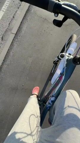 A first-person perspective video shows a person riding a bicycle on a paved path. The rider's legs, wearing light-colored sweatpants and pink clogs, are visible in the foreground. The bicycle's handlebars, brakes, and a digital display are also in view. The background shows a curb, a road with a white line, and a grassy area with trees and a dark fence. The sun casts shadows on the path. The rider briefly turns the camera towards their face, a young woman with brown hair pulled back, who smiles and laughs as she rides. The video captures a moment of outdoor activity and enjoyment.