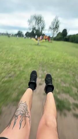 The video shows a first-person perspective of someone on a swing set in a park. In the foreground, the person's legs and black shorts are visible. The swing is moving, causing a blurred effect in the video. In the background, a grassy field with playground equipment like a slide and seesaw can be seen. There are a few people, including children, playing in the park. The sky is cloudy. A young girl in a purple shirt is visible on a swing in the right side of the frame.