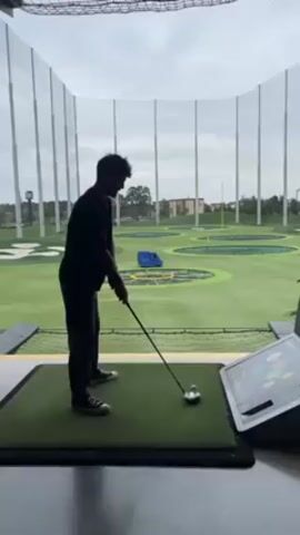 The video shows a man playing golf at Topgolf. The man is standing on a green mat, holding a golf club, and preparing to swing at a golf ball. The background shows a large, open area with several targets and nets. The man swings the club and hits the ball, and then stops.