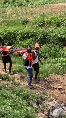 The video shows a group of four people carrying a person on a red stretcher through a rough, overgrown area. The person on the stretcher appears to be male and is lying still. The other three individuals seem to be helping carry the stretcher through the uneven terrain, surrounded by lush green foliage and some scattered trash. One person in the group wears an orange life vest and appears to be talking on a phone. The environment looks like a natural area mixed with signs of human activity and waste.