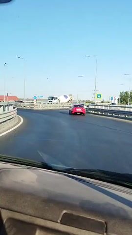 The video shows a view from inside a vehicle traveling on a road. In the distance, a concrete mixer truck is visible on a roundabout. A red car is also seen in front of the truck. The sky is clear and blue, indicating daylight. The road appears to be asphalt, and there are streetlights along the side. The perspective suggests the vehicle is in motion, possibly navigating the roundabout.