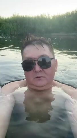 A man wearing sunglasses is submerged in water, with his upper body visible. He is looking at the camera, and the water is reflecting the sky and trees. The background consists of lush green foliage.
