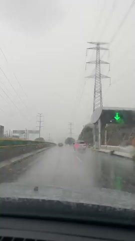 The video shows a view from inside a vehicle traveling on a wet road during what appears to be daytime and overcast weather. Rain is visible on the windshield, and the road surface is slick with water. Several other vehicles are on the road ahead, including a white SUV. Tall electrical pylons are visible in the background, along with power lines stretching across the sky. There is a structure resembling a toll booth or entry gate on the right side of the road, with a green arrow sign illuminated. The overall impression is one of driving in inclement weather.