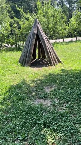 This is a vertical video of a rustic, conical structure made of rough-hewn logs, resembling a primitive hut or shelter. The structure is set in a grassy, outdoor environment with green trees in the background and more grass and clover in the foreground. The sun is shining, casting shadows from the logs onto the ground and creating dappled light on the surrounding grass. The time of day appears to be daytime, likely late morning or afternoon, given the brightness and the angle of the shadows. The setting suggests a park, a garden, or a natural area in Tesserete, Switzerland. There are no people visible in the frame, and the focus is solely on the wooden structure and its natural surroundings. The overall impression is one of simplicity and nature.