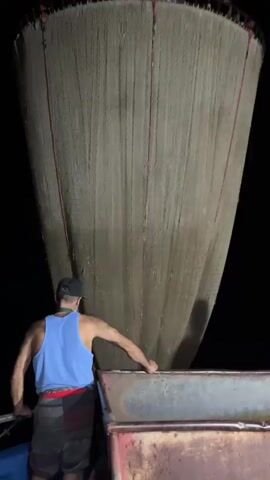 A person wearing a blue tank top, black baseball cap, and plaid shorts stands on a boat at night, holding a large fishing net. The net, made of light-colored mesh with red vertical stripes, hangs down from the top of the frame. The person is pulling on the net, which is funnel-shaped and appears to be filled with something. The boat has a metal railing. The background is dark, suggesting it is nighttime and the boat is on the water.
