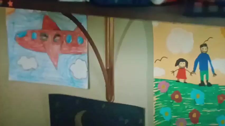 The video pans across a wall displaying children's artwork and various craft supplies. On the left, a red airplane with blue windows is depicted against a cloudy sky. Next to it, a drawing shows a smiling man and a girl holding hands in a field of colorful flowers under a yellow sky with birds and a sun. Below these, a dark drawing with a crescent moon and stars is visible. To the right, a shelf holds bottles of glue and other craft items, including a blue basket containing a roll of tape and what appears to be a marker. A green toy dinosaur is placed in front of a drawing of a boy with a green shirt holding a radio in front of what looks like a house. To the right of this drawing is a picture of a sunrise over water with pink clouds. The overall environment appears to be a child's creative space.