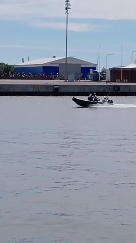 A black rigid inflatable boat is traveling from left to right across the screen. Two people are visible inside the boat, one at the helm and one standing. The boat is creating a wake. In the background, there is a concrete pier with several buildings and structures, including a tall white pole with lights. A line of people are visible standing on the pier. The sky is blue with some clouds. The water is choppy.