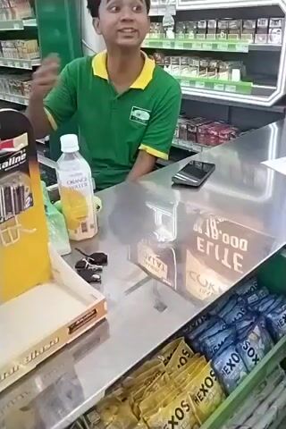 A young man wearing a green shirt with a yellow collar is standing behind a counter in what appears to be a convenience store. He is looking at the camera and speaking. On the counter, there are several items, including a bottle of orange water, a black smartphone, a set of keys, and a display of batteries. In front of the counter, there are rows of packaged goods, possibly condoms, in various colors. The background shows shelves stocked with products, likely cigarettes and other convenience store items. The man gestures with his hands as he speaks.