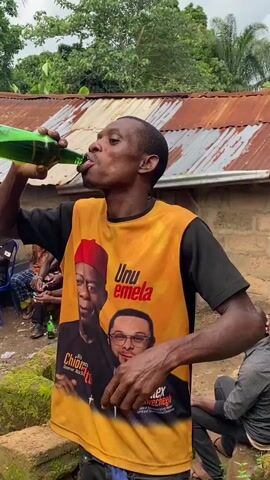 A man wearing a yellow and black t-shirt with political images and text is drinking from a green glass bottle. The t-shirt has two faces on it, one with a red hat and the other wearing glasses. The man is holding the bottle to his mouth and tilting his head back as he drinks. He then lowers the bottle, wipes his mouth with his hand, and appears to be speaking. In the background, other people are visible sitting on chairs, and there is a corrugated metal roof and some greenery. The scene appears to be outdoors in a rural setting.