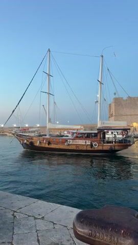 The video shows a harbor scene at dusk. A large wooden sailboat with two masts is docked in the foreground, its reflection visible in the calm water. In the background, there is a stone fortification or building, and further off, more boats and harbor structures can be seen. The sky is a clear blue with a hint of sunset colors near the horizon. People are visible sitting on benches along the waterfront and walking in the distance. A black car is parked on the cobblestone walkway. The video pans slightly, offering different views of the harbor and the boats.