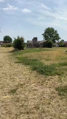 The video pans across a dry, grassy field with some trees and structures in the background. A narrow path is visible, leading towards the structures. The sky is cloudy and blue. In the distance, there appear to be some buildings or ruins, and a few trees are scattered across the landscape. The camera continues to move, showing more of the dry grass and vegetation. There is a small stream or body of water visible in the latter part of the video, with green grass and bushes lining its banks. Further in the distance, there seems to be a fence or netting, possibly related to a sports field. The overall impression is of an outdoor, somewhat overgrown and neglected area on a sunny day.