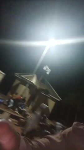 The video, shot at night under an artificial light, appears to be a shaky first-person perspective. The camera is angled upwards initially, showing a cluttered scene with boxes and other indiscernible objects. The camera then pans downwards, revealing a person, presumably the one holding the camera, lying on the ground. It's dark and difficult to make out details, but the person is wearing a light-colored shirt. A bicycle is also visible nearby. The environment seems to be outdoors, possibly a grassy area.