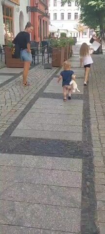 A woman in a black shirt and denim shorts walks on a cobblestone path, holding a black bag. She is followed by two young girls, one in a white t-shirt and black shorts, and another in a blue t-shirt and tan shorts, carrying a stuffed animal. They are walking away from the camera down a street lined with buildings and planters. The street is paved with cobblestones and large stone slabs.