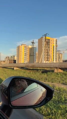 A car is driving past new apartment buildings. The apartment buildings are tall and colorful, with yellow, beige, and white facades. They appear to be under construction or recently completed. In the foreground, a car's side mirror reflects the interior of the car and the person driving. Beyond the buildings, there are power lines and poles. The environment is grassy with a large pipe running horizontally in the middle ground. The sky is clear and blue, suggesting it is daytime.