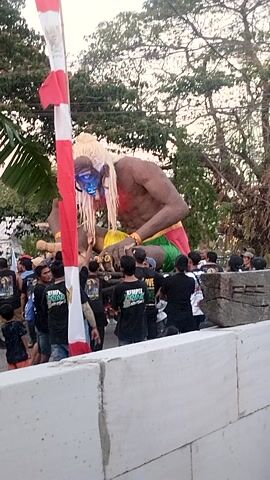 A vertical, slightly shaky, full shot captures a street celebration with a large, inflatable figure as the focal point. The figure, resembling a giant with muscular arms and legs, is adorned with red face paint, blue eyes, and white hair. It wears a green and yellow garment around its waist and a yellow necklace. In front of the giant, a red and white striped banner stretches across the scene, with a banana leaf positioned on the left. A crowd of people, mostly wearing black t-shirts with various designs, are gathered around the figure, some looking at it, others talking amongst themselves. They appear to be observing or participating in the event. The environment suggests an outdoor setting with trees visible in the background and what appears to be a white concrete structure on the right side of the frame. The lighting indicates it is likely late afternoon or early evening. The overall atmosphere seems festive and communal.