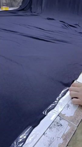 A pair of hands is shown cutting a large piece of navy-blue fabric with a pair of black and silver scissors. The fabric is laid out on what appears to be a large, tiled table. There is clear plastic on the edge of the table. The person cutting the fabric is wearing a gray t-shirt. The hands have some writing on them. In the background, more of the same fabric is draped. The background is out of focus.  The action consists of cutting the fabric. The environment is a large room with a tiled table. The setting appears to be a workshop or factory.