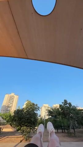 The video shows a first-person perspective of someone sitting in a park with their legs outstretched. The person is wearing black shorts and white sneakers. In the background, there are trees, benches, and tall residential buildings, indicative of an urban park setting in Givat Shmuel, Israel. The sky is a clear, bright blue, suggesting it's daytime, likely late afternoon due to the warm lighting and long shadows visible on the ground. The person appears relaxed, enjoying the serene atmosphere of the park. There is a large, beige canopy providing shade over the viewer. The ground beneath the viewer has a colorful, textured surface with red and tan sections. Another person is visible sitting on a bench in the distance. No visible text is present in the video.
