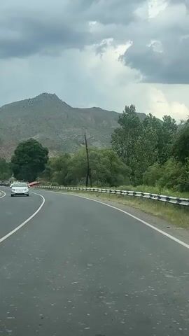 The video captures a forward-facing view from a moving vehicle driving along a winding asphalt road in a rural, mountainous setting. The road has clear white lane markings and features a white metal guardrail with blue accents on the right side. Green trees and dense bushes line both sides of the road, and a tall wooden utility pole is visible on the right. In the background, prominent mountains with sparse vegetation and rocky peaks rise under an overcast, cloudy sky. The road surface shows scattered wet patches, suggesting recent rain.

At the beginning of the clip, a white sedan is seen ahead, navigating the curve. As the vehicle progresses, other cars become visible, including a red semi-truck driving in the opposite direction around the 0:02 mark. Further down the road, several white cars are parked on the right shoulder near a small, partially visible building or structure. A blue and white road sign is also faintly discernible in the distance. The scene portrays a calm, daytime journey through a scenic, green landscape, consistent with the region of Chiva, Armenia. No readable text is visible on any signs or objects.