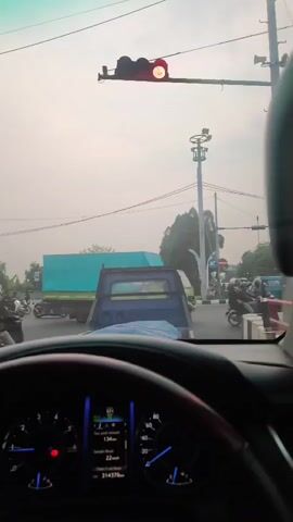 This is a dashcam video from inside a vehicle stopped at a traffic light. The traffic light is red. In front of the vehicle, there are several other vehicles including trucks, motorcycles, and cars. The environment appears to be an urban or suburban area with traffic infrastructure such as traffic lights and utility poles. The sky is overcast. The dashboard of the vehicle is visible in the foreground, showing the speedometer, fuel gauge, and other indicators. The video shows a short period of time where the vehicles are stationary due to the red traffic light.