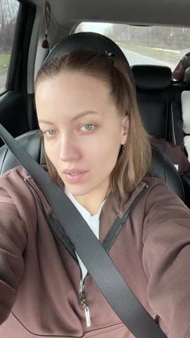 A young woman with light brown, shoulder-length hair and light-colored eyes is seated in the passenger seat of a car, recording a selfie video. She is wearing a brown zip-up hoodie over a white top, with a seatbelt across her chest. She is speaking and singing along to music playing in the background, appearing relaxed and happy.

The car's interior is visible, including a car seat in the back. A small decorative charm hangs from the rearview mirror. Through the car windows, a moving landscape of trees and foliage suggests the car is in motion on a road. The weather outside appears overcast.

At the 0:02 mark, the video rotates 90 degrees clockwise, showing the woman's face from a slightly different angle. In the background, on items in the back seat, the words "Junior" and "SONAME" are visible.
