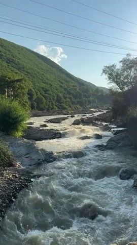 The video shows a wide, fast-flowing river in a mountainous region. The river is rocky with rapids and white water. The banks of the river are covered in lush green vegetation, and a steep, tree-covered mountain rises on the left side of the frame. The sky is clear and blue, with a few wispy clouds. Power lines are visible strung across the sky in the upper part of the frame. The sun appears to be shining, casting light on the water and the landscape. The overall impression is of a wild and natural environment.