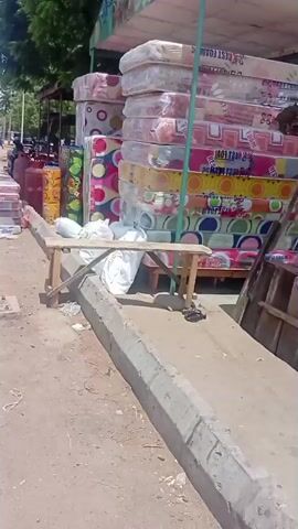 The video captures an outdoor commercial scene on a sunny day, panning slowly from right to left. Multiple stacks of colorful mattresses, individually wrapped in clear plastic, are displayed along a dirt path next to a concrete curb. White bags rest on the curb. As the camera pans, more mattresses, some stacked horizontally and others rolled vertically, are visible. Large red gas cylinders are positioned among the goods. In the mid-background, a man wearing a light-colored robe walks from left to right. Several motorcycles are parked further back near a building with corrugated metal siding, partially painted with yellow, blue, and red stripes. The pan concludes with a view of the stacked mattresses, gas cylinders, and rolled items in front of the striped building section.