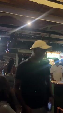 The video shows a dimly lit outdoor space, possibly a patio or street corner, crowded with people. The crowd is diverse, with individuals of varying skin tones and styles of dress. Most people are standing and appear to be engaged in conversation or enjoying music, as some are dancing and raising their arms. The background includes signs like "M/S. SHRIDHAR WAS," suggestive of nearby businesses or establishments. Christmas-themed string lights and star decorations hang overhead, adding to the festive atmosphere. A few wooden tables and chairs are visible in the foreground, hinting at a casual outdoor dining or gathering area.