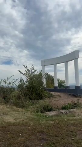 A video taken in landscape orientation, shows a white architectural structure with three columns and a curved top piece, standing on a hilltop. The structure is positioned slightly to the right of the frame, with a large bush to its left. The ground in the foreground is covered with dry grass and some patches of dirt. Beyond the structure, a panoramic view of a valley can be seen, with a body of water and distant hills. The sky is filled with dramatic, fast-moving clouds, suggesting a dynamic weather system. The lighting indicates it is daytime, likely late afternoon or early evening, given the subdued sunlight filtering through the clouds. There are no people visible in the video.