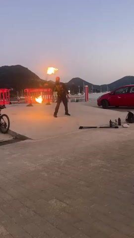 A person stands in the middle of a paved area, holding two flaming objects, one in each hand, and appears to be spinning them. The flames are bright orange and cast a warm glow. The person is wearing dark clothing. In the background, there are red barriers, a red car, and hills with trees. The sky is a dusky blue, indicating it is either dawn or dusk. There is a bicycle on the left side of the frame, and some equipment, possibly related to the fire performance, is on the right side of the frame.