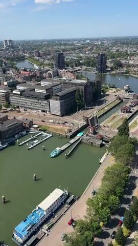 The video shows a canal with two barges and a bridge. The barges are green and white and are docked in the canal. The bridge is a drawbridge and is open, allowing the barges to pass through. There are buildings and trees in the background. There are cars and people on the street. The weather is sunny.