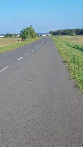 A drone flies over a road in a rural area. The drone is flying at a relatively low altitude, and it is possible to see the details of the road and the surrounding landscape. The road is paved with asphalt, and there are white lines painted on the road to mark the lanes. The surrounding landscape is fields of grass and trees. The sky is blue and there are a few clouds. The drone is flying in a straight line, and it appears to be moving at a constant speed.