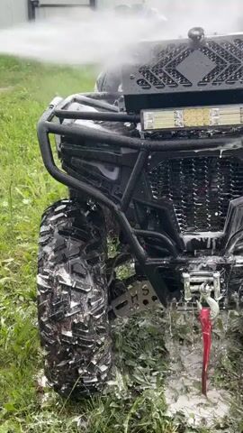 A black all-terrain vehicle (ATV) with a large knobby front tire, brush guard, light bar, and winch is being sprayed with water and white foam. The pressure washer is off-screen to the left, and a stream of water is visible hitting the front left side of the ATV. Foamy white suds are dripping down the vehicle onto the green grassy ground.