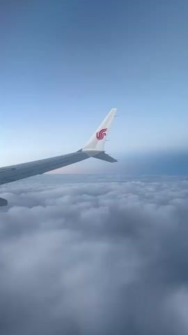This is a point-of-view shot from an airplane window. The left side of the frame shows the wing of the airplane, with the China Southern Airlines logo visible on the wingtip. The wing extends towards the left edge of the frame. Below the wing is a vast expanse of white clouds, covering the entire bottom half of the image. The sky above the clouds is a clear, pale blue, transitioning to a darker blue on the right side of the frame, suggesting the presence of the horizon or a change in atmospheric conditions. The lighting is soft, indicating either sunrise or sunset. There are no people visible. The camera is steady, suggesting the airplane is in smooth flight.