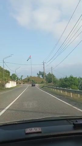 The video shows a road scene from inside a vehicle. A large flag on a tall flagpole is visible on a hill to the right of the road. The flag is red at the top and has alternating green and white stripes below, with a white star and crescent moon symbol in the red section. A dark-colored car is driving on the road ahead of the camera. There are trees and bushes along the sides of the road and on the hill. Power lines are visible in the sky. The sky is blue with some clouds. There is a bus stop shelter near the base of the hill. The camera appears to be moving forward along the road.