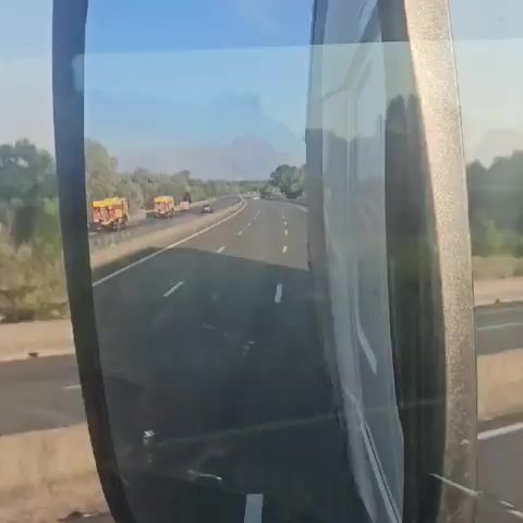 The video shows a highway scene. On the left side of the highway, there are several yellow and red trucks that appear to be fire trucks. They are driving in a convoy. There are also some trees and vegetation on the left side of the road. In the distance, there is smoke rising, suggesting a fire. On the right side of the highway, there is a reflection in a mirror, possibly from a vehicle. The sky is blue with some hazy clouds.