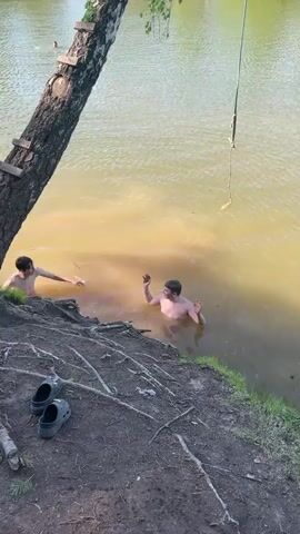 A daytime full-shot shows two young men swimming in a murky, brown-green lake under a clear, blue sky. The men stand on a shallow bank, submerged from the chest down in the lake. The man on the right, with a bare torso, stands upright and smiles, gesturing with his hands. The man on the left faces the bank, with only his head and shoulders visible. In the foreground, a large tree with thick roots extends from the left bank. A rope swing hangs over the water from a tree branch on the right. In the background, a person swims in the distance, and the opposite bank is covered with dense green trees. The lake water transitions from a murky brown near the bank to a clearer blue-green further out.