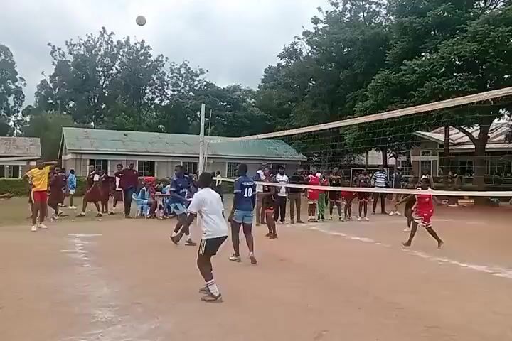 The video shows a volleyball game taking place outdoors. Two teams of young people in different colored uniforms are playing on a sandy court with a net dividing the two sides. The team on the left is wearing blue and white, while the team on the right is in red and black. There are several spectators watching the game, including people in casual clothing and some in school uniforms. The background consists of buildings, trees, and a cloudy sky. The video captures the players' movements as they hit and pass the volleyball during the game.
