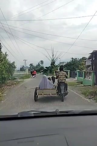 The video captures a view from a vehicle looking down a paved road. In the foreground, the dashboard of the vehicle is visible. The road is lined with vegetation and houses. In the middle ground, a motorcycle with a sidecar is traveling away from the camera. The motorcycle is driven by a person wearing a patterned shirt. Inside the sidecar, three individuals are seated. One person is wearing a light-colored head covering, and two children are also visible. Further down the road, a group of four people are walking away from the camera. The sky above is overcast with visible power lines crisscrossing the frame. The environment appears to be rural or suburban.