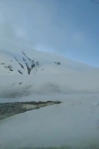 The video displays a snow-covered mountainous landscape under a partly cloudy sky, as seen from a moving vehicle. A vast expanse of white snow blankets the mountain slopes, with some darker patches of rock or earth visible in places where the snow cover is thinner. A road runs along the bottom of the frame, with snow piled up on its sides. At one point, a red and white chevron road sign, indicating a sharp curve, is visible on the side of the road, partially covered in snow. A simple metal fence runs alongside another section of the road. The scene suggests a winter or early spring journey through a high-altitude region, specifically Ganisi, Georgia. No people are visible in the video.